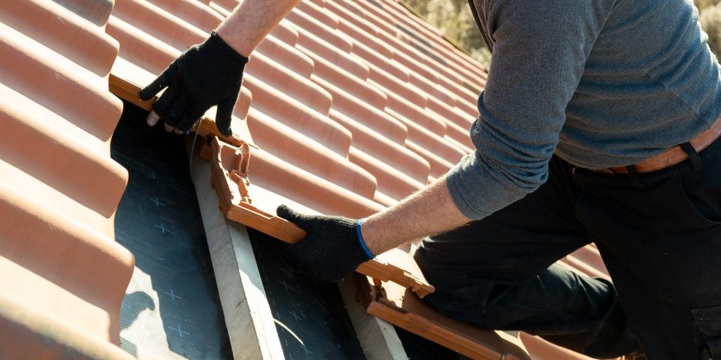 Closeup of worker hands installing yellow ceramic roofing tiles mounted on wooden boards covering residential building roof under construction.
